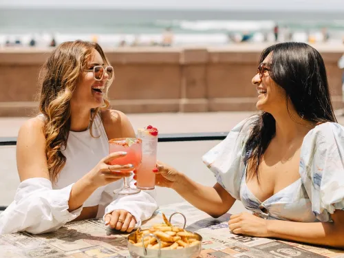 Two women clinking glasses with drinks at a beachside table at Belmont Park in San Diego, smiling and enjoying the moment.