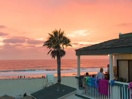 Sunset over the ocean at the Beach Cottages in San Diego, with vibrant pink clouds, palm tree, and beachgoers in the distance.