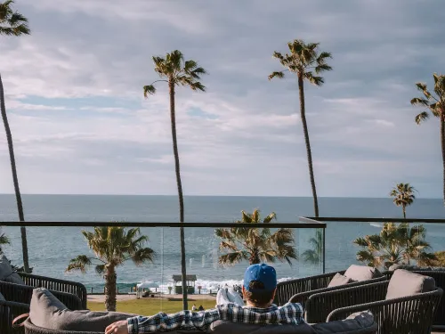 A person relaxes on a couch, overlooking the ocean and palm trees under a cloudy sky at the La Jolla Cove Hotel in San Diego.