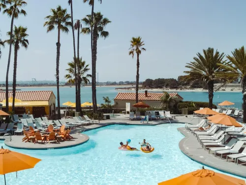 Loungers and umbrellas by a pool at the San Diego Mission Bay Resortwith palm trees and a scenic beachfront view.