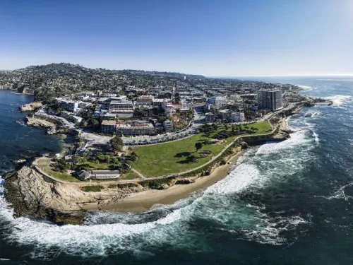 Coastal view of a vibrant town with greenery, cliffs, and ocean waves under bright sunlight.