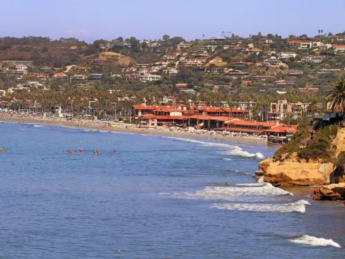 Coastal view of the La Jolla Shores Hotel in San Diego, featuring sandy beach, waves, and palm-lined hillside with colorful buildings.