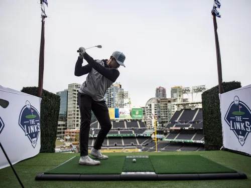 A golfer swings a club on an artificial green with the San Diego skyline in the background.