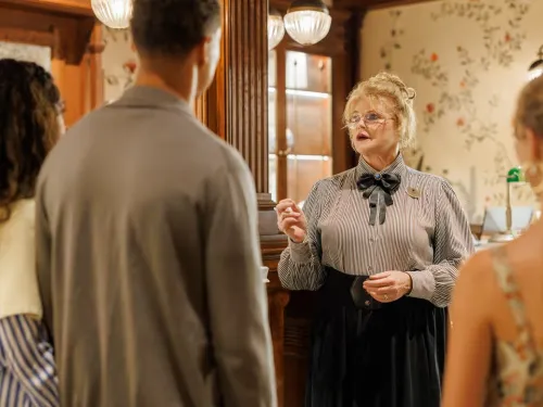 A blonde woman in period attire speaks to a group of three people in a cozy room as part of the Hotel del Coronado's Haunted Happenings Tour near San Diego.