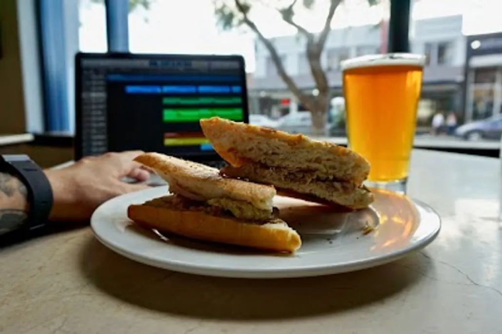 A plate with a sandwich, a glass of beer, and a person using a laptop on a table in a cafe with a street view outside the window.
