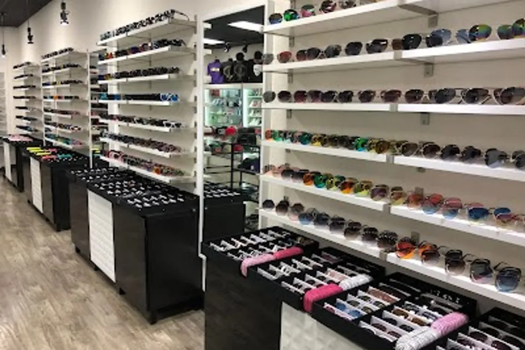 Rows of sunglasses are displayed on white shelves and black tables in a well-lit retail store with a wood floor.