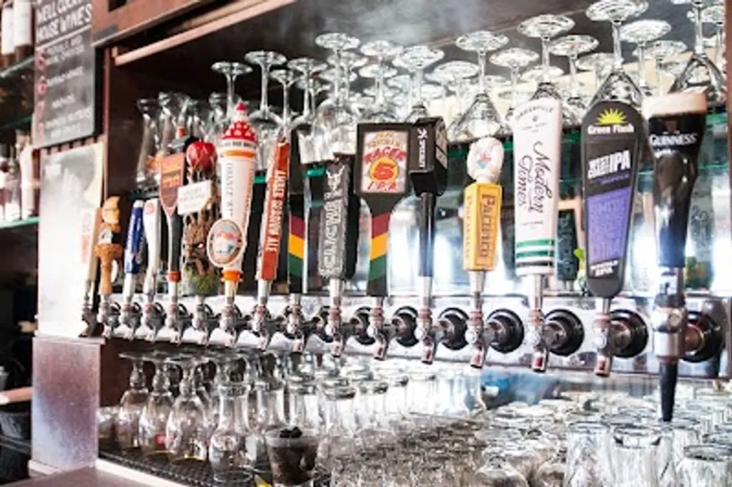 A row of beer taps with various branded handles is mounted above a bar counter with empty glassware hanging and stacked below.