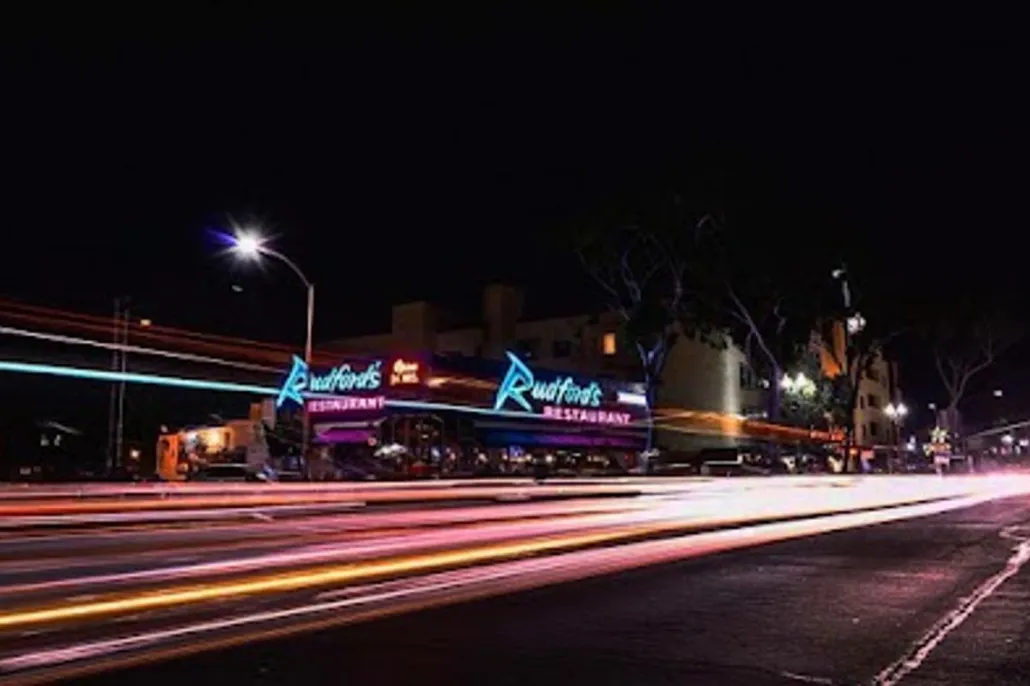 Nighttime street scene with light trails from passing cars in front of a neon-lit restaurant named “Rudford’s Restaurant.” Trees and buildings are visible in the background.