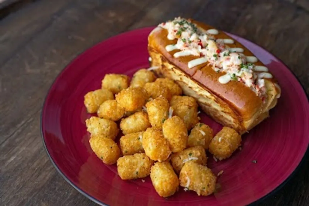 A red plate with a lobster roll topped with white sauce and herbs, served alongside a pile of crispy tater tots.