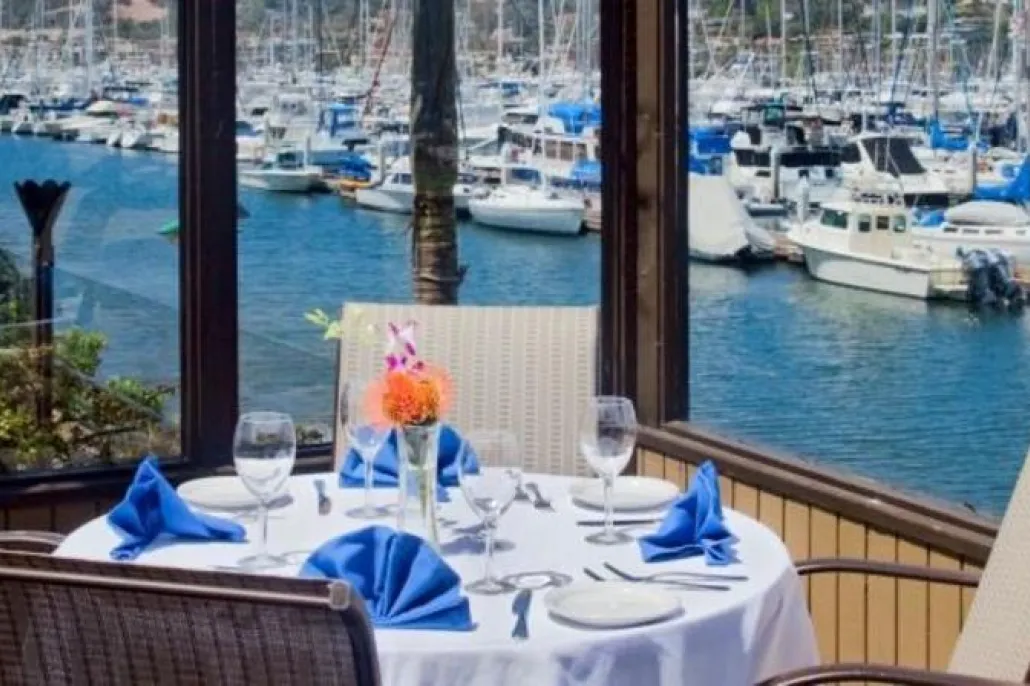Table set for four with blue napkins and glassware, overlooking a marina filled with docked boats through large windows.