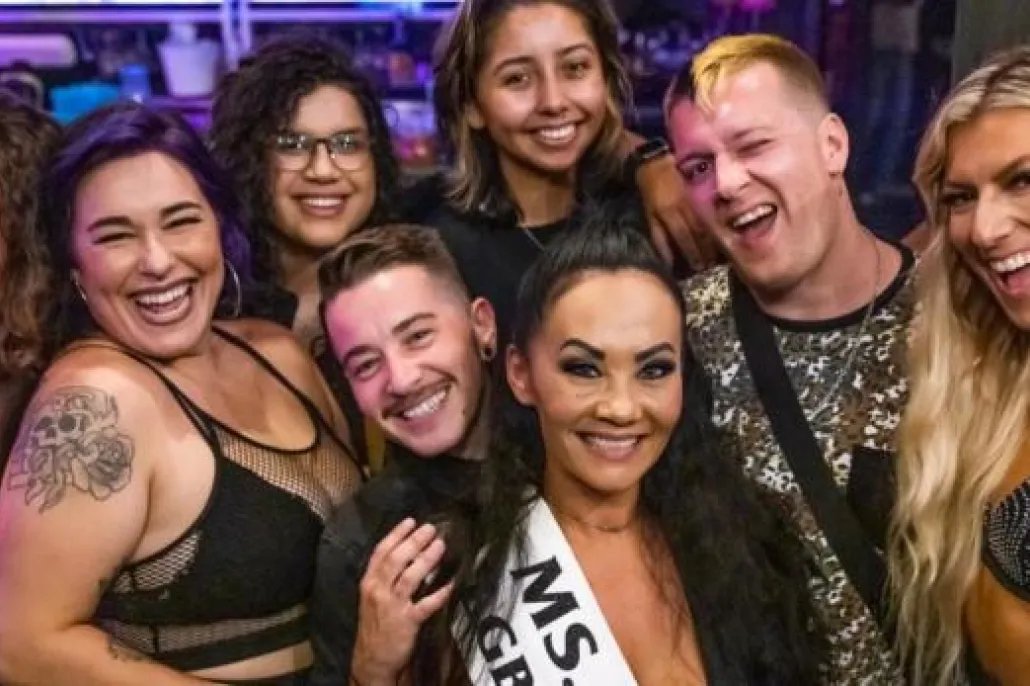 A group of eight people, some smiling and some posing, stand close together indoors at what appears to be a lively event. One person wears a "MS." sash.