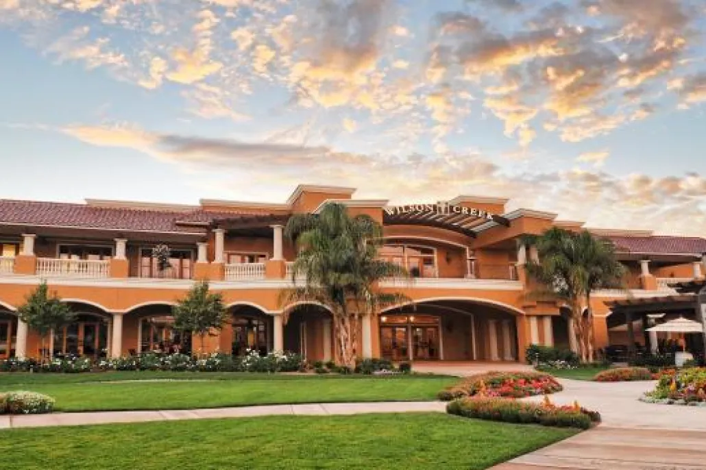 A large, elegant building with arched entrances and balconies sits amid manicured lawns and palm trees under a sky with scattered clouds at sunset.