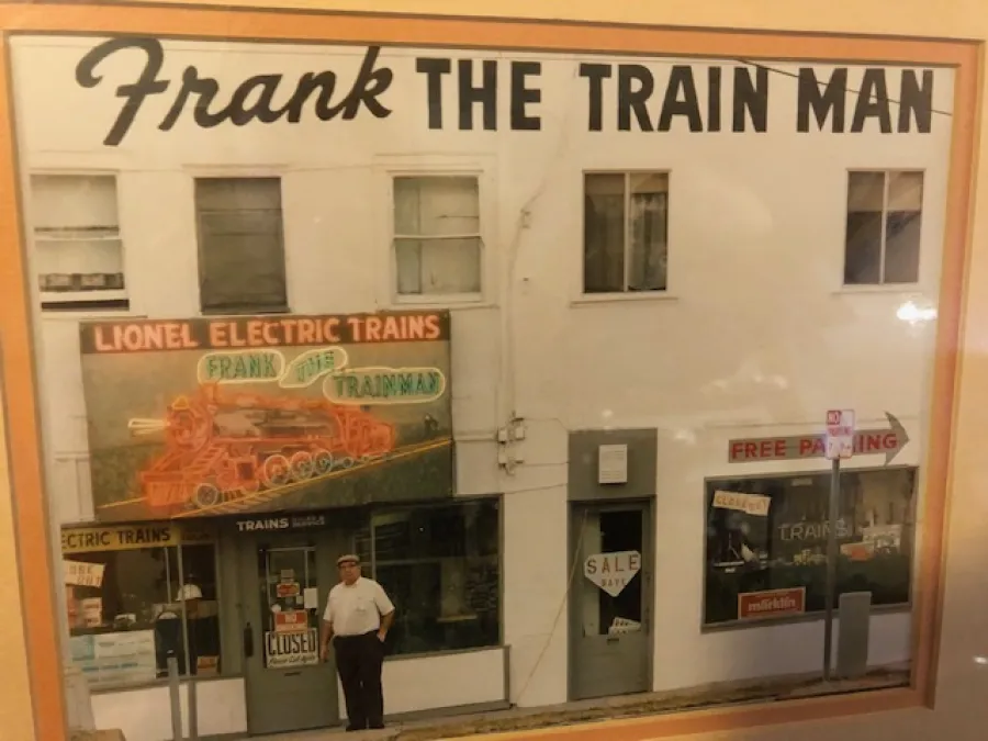 A man stands in front of a store called "Frank the Train Man," featuring a neon sign for Lionel Electric Trains and a "Closed" sign on the door.