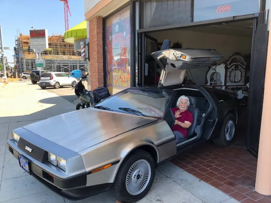 An older woman sits smiling inside a silver DeLorean with gull-wing doors open, parked outside a storefront on a sunny day.