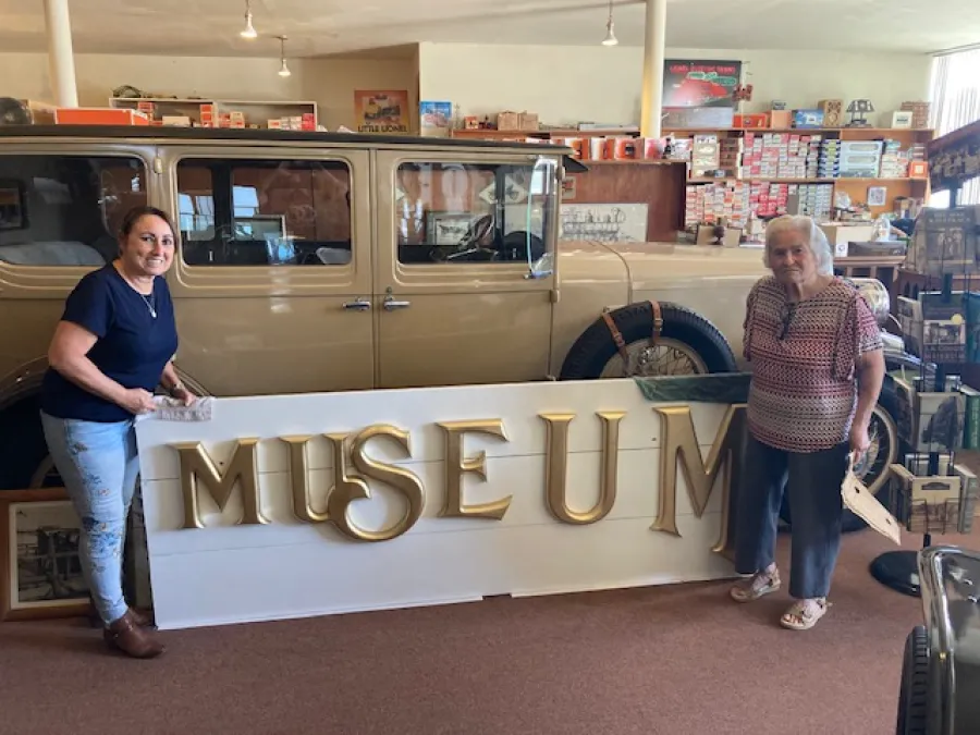 Two women stand next to a vintage car, holding up large gold letters spelling "MUSEUM" inside a room filled with various antiques and collectibles.
