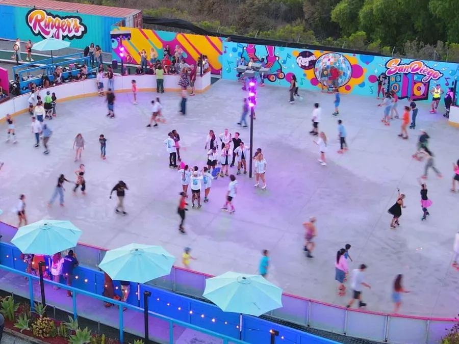 Aerial view of Ringer’s Roller Rink in Encanto, San Diego, with skaters gliding across a large outdoor rink surrounded by colorful murals, string lights, umbrellas, and spectators enjoying a lively evening atmosphere.