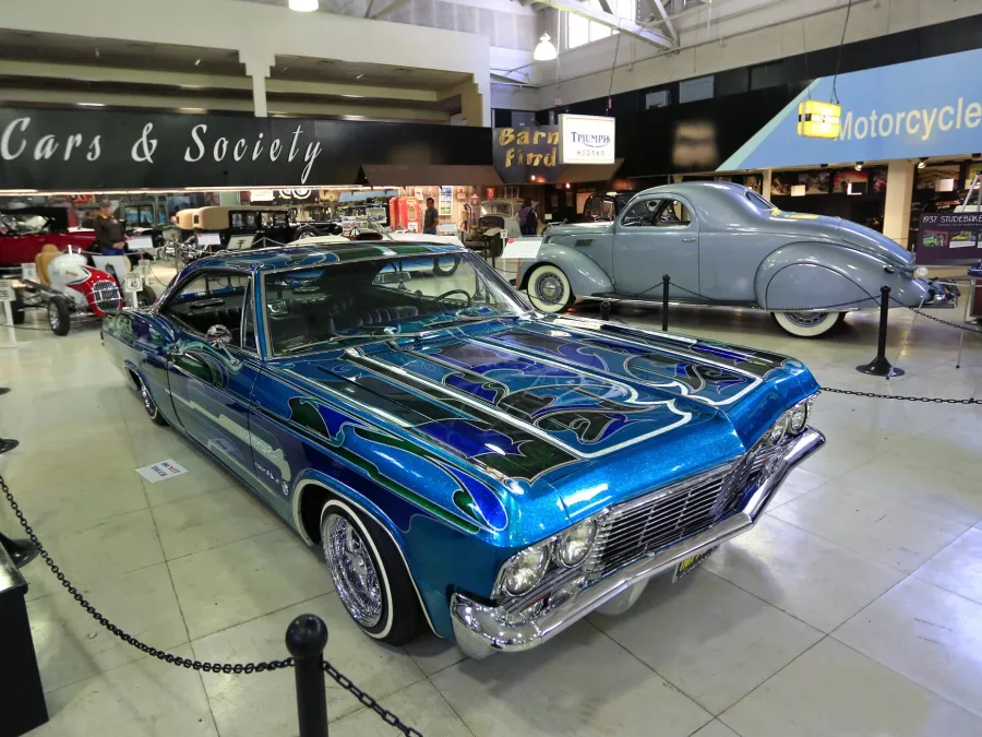 A sleek blue racing car sits in the foreground among other vehicles at the San Diego Automotive Museum.