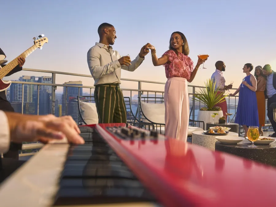 A lively rooftop party scene at Mister A's in San Diego, with people dancing, playing instruments, and enjoying drinks at sunset.