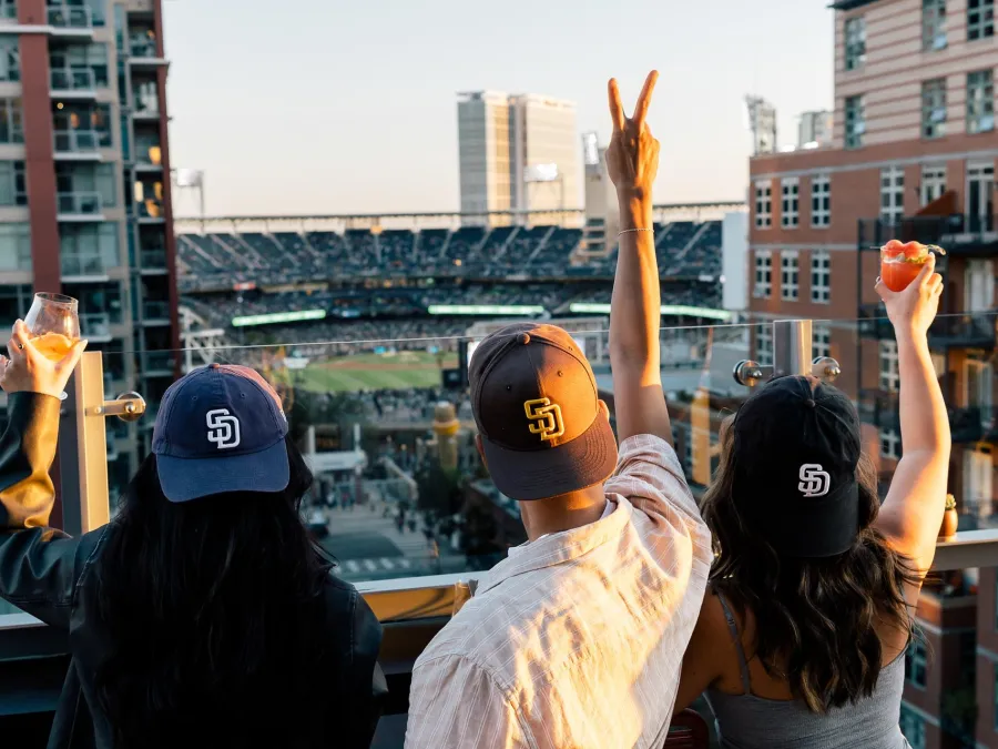 Three friends in baseball caps raise drinks at Borrego, overlooking a Petco Park at sunset.