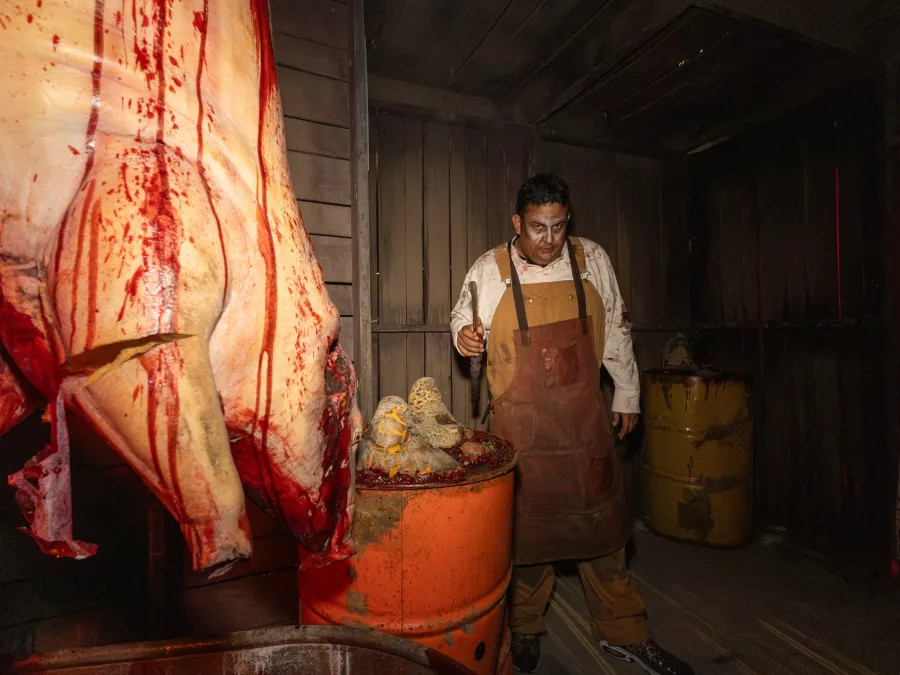 A person in a bloody apron stands in a dimly lit room with hanging meat in one of SeaWorld San Diego's Howl-O-Scream Halloween haunted houses.