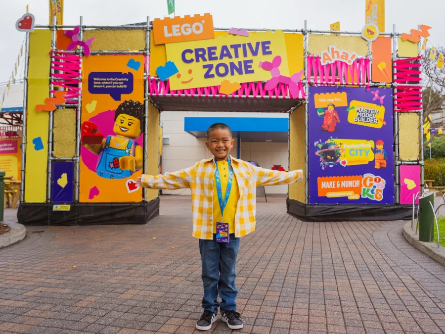 A child in a yellow checkered shirt smiles with arms outstretched in front of a colorful LEGO entrance of one of the LEGO Festival play zones at LEGOLAND California.