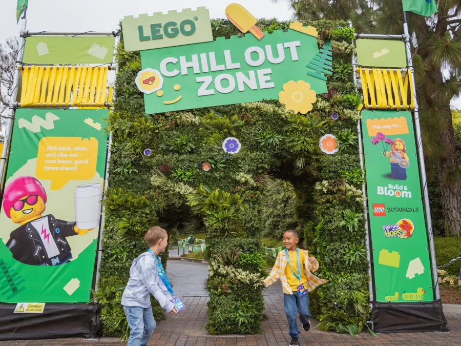 Two children play at the entrance of a vibrant LEGO-themed "Chill Out Zone" filled with greenery at LEGOLAND California.