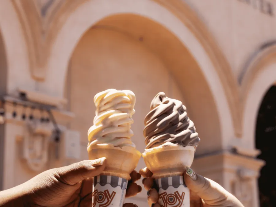 Two hands holding ice cream cones from Element 8 Cafe in front of The Fleet Science Center with arches.