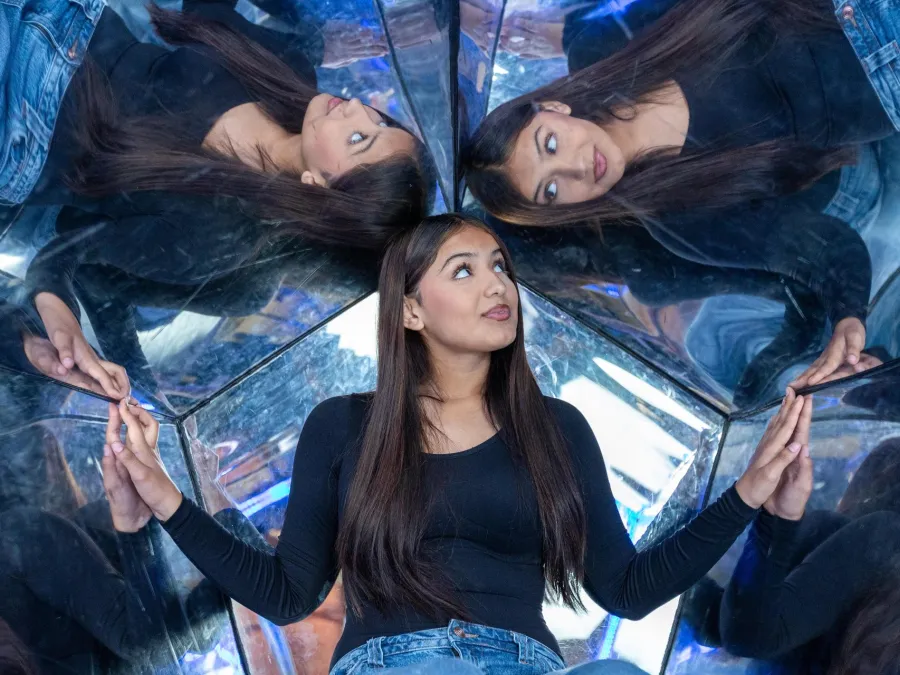 A woman poses with outstretched hands in a mirrored setting, reflecting multiple images of herself at the Fleet Science Center in Balboa Park.