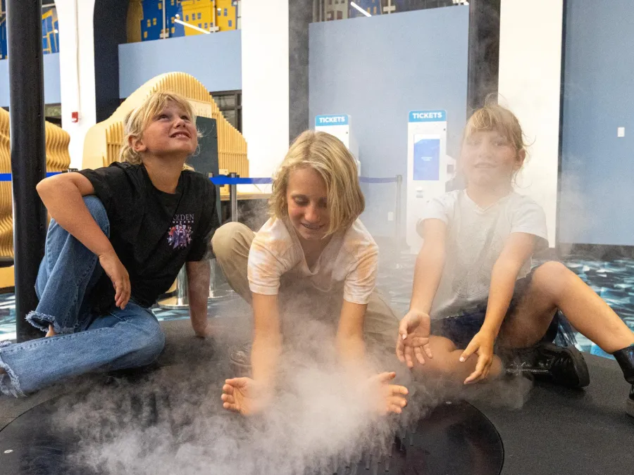 Three children sitting on a floor covered with mist, smiling and playing together at the Fleet Science Center in Balboa Park.
