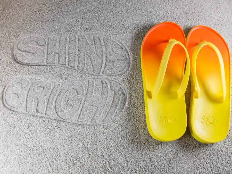 Yellow and orange flip-flops beside the impression of "SHINE BRIGHT" in sand.
