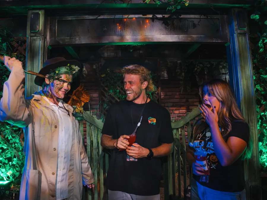 A smiling man and woman interact with a costumed character in a vibrant, illuminated setting  at SeaWorld San Diego's Howl-O-Scream Halloween event.