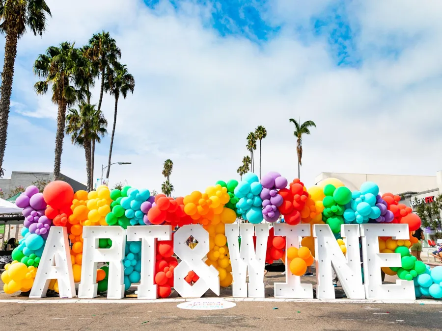 Colorful balloon arch with the words "ART & WINE", surrounded by palm trees and a blue sky at the La Jolla Art & Wine Festival in San Diego.