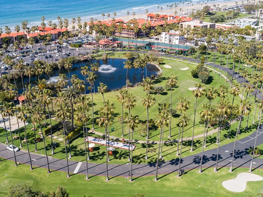 Aerial view of a circular park at La Jolla Beach & Tennis Club, with palm trees and a pond, near a beachside resort.