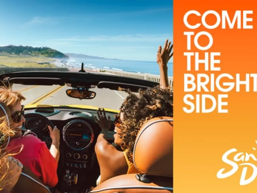 Three people drive a convertible along a coastal road with the ocean visible, next to orange text reading "Come to the Bright Side, San Diego" with a sun graphic.