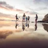 A group of people stand and play with a ball on a wet sandy beach at sunset, with cliffs and ocean visible in the background.