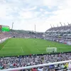 A large soccer stadium filled with spectators shows a match in progress on a green field under a partly cloudy sky.