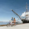 A couple riding a bike on San Diego's Embarcadero with the Star of India at the Maritime Museum in the background