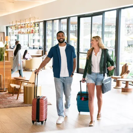 A man and woman walk through the stylish hotel lobby of the Town and Country Resort, each with a suitcase, smiling.