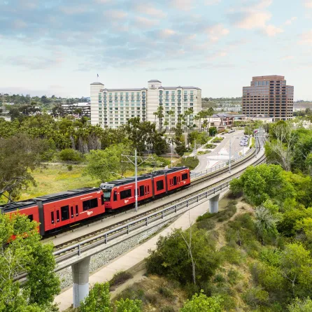 San Diego Red Trolley on a railway surrounded by greenery, buildings, and a cloudy sky.
