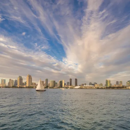 San Diego skyline at sunset with dramatic clouds and a sailboat in the foreground on calm water.