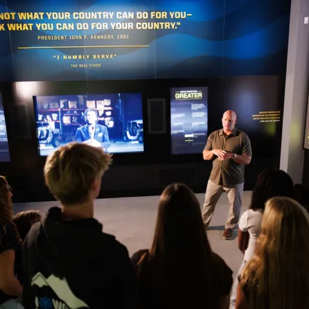 A group of people listens to a speaker in an exhibit with screens and text on the wall at the Navy SEAL Museum San Diego.
